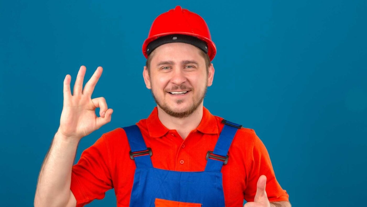 Builder Man Wearing Construction Uniform And Security Helmet Smiling Friendly Doing Ok Sign And Showing Thumb Up Over Isolated Blue Wall