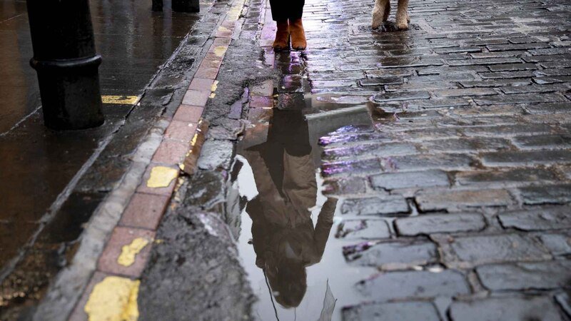 Mature Woman Walking Her Dog In The City Streets While It Rains Water Pooling At End Of Driveway