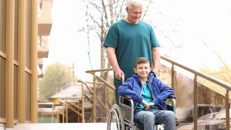 Preteen Boy In Wheelchair With His Grandfather Using Ramp Outdoors